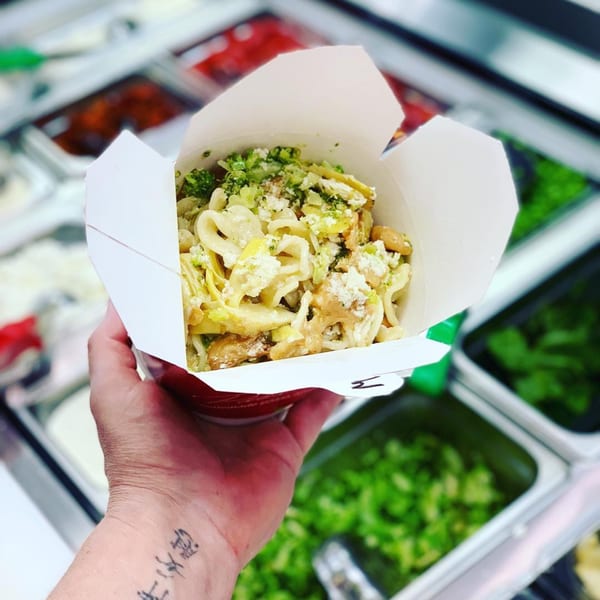 Hand holding a Pasta Volo takeout box filled with pasta, broccoli, and cheese, above ingredient bins.