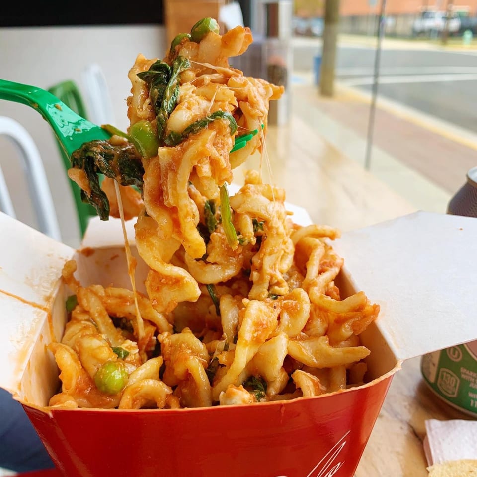 Cheesy pasta with peas and spinach being lifted from a red takeout box with a green fork.