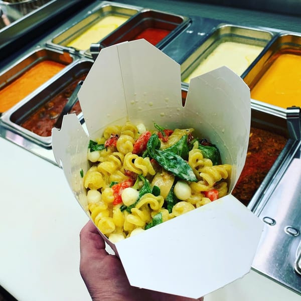 A hand holds a white takeout box filled with Pasta Volo's pasta, mozzarella, tomatoes, and basil.