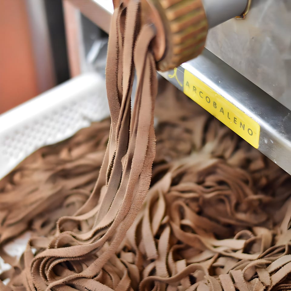 Freshly extruded brown pasta, possibly chocolate or whole wheat, emerging from a pasta machine.