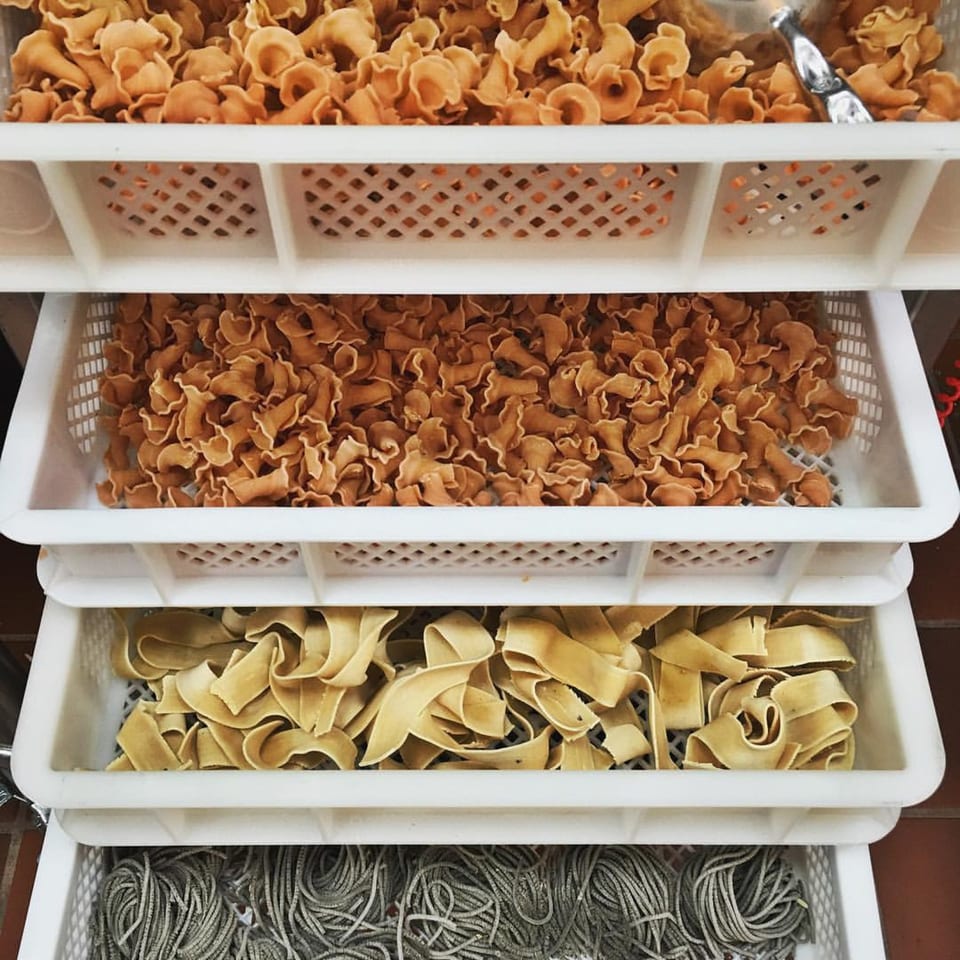 Four white trays stacked with different types of fresh, uncooked pasta, including campanelle, pappardelle, and spaghetti.