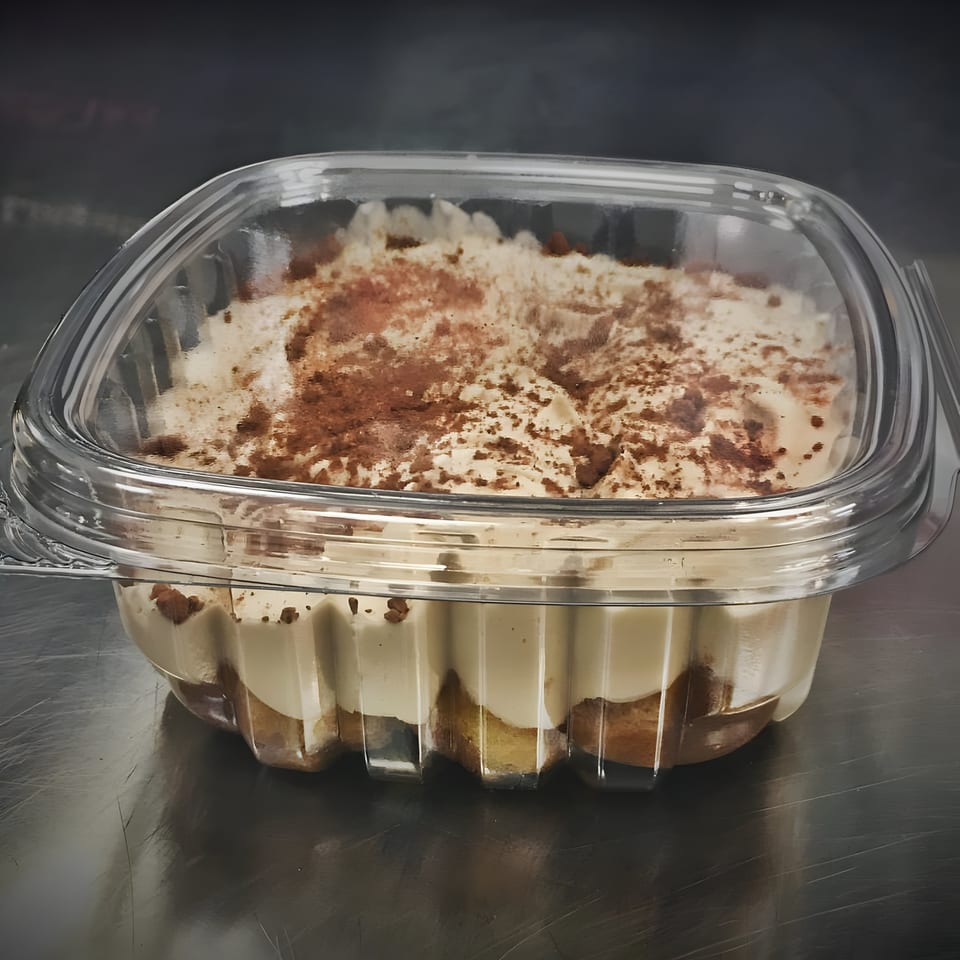 Tiramisu dessert in a clear plastic container, topped with cocoa powder, on a dark metallic surface.