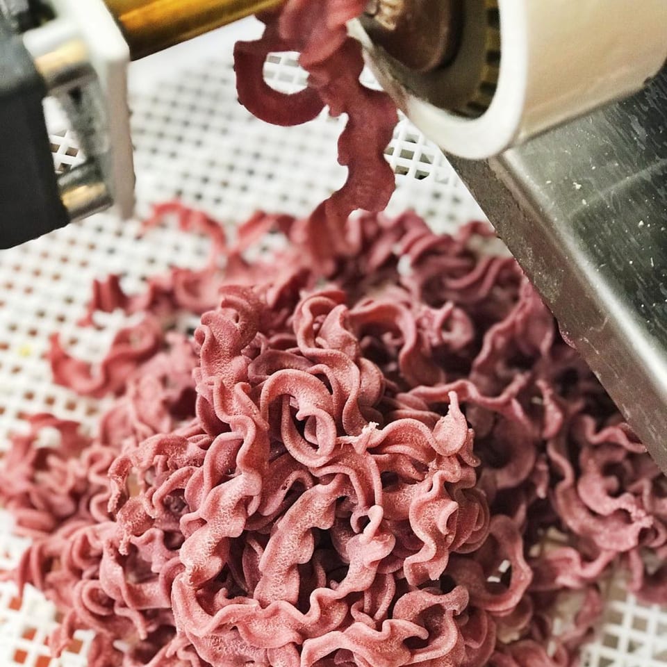 Freshly extruded red pasta, possibly beet-infused, curls onto a white drying rack from a pasta machine.